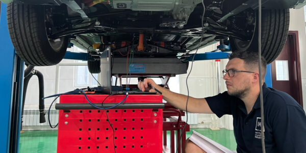 A trainer (Pete Banks), researching an EV200 BAIC electric vehicle on behalf of a client. He's using a laptop and is working underneath a vehicle raised up on a 2 poster ramp.